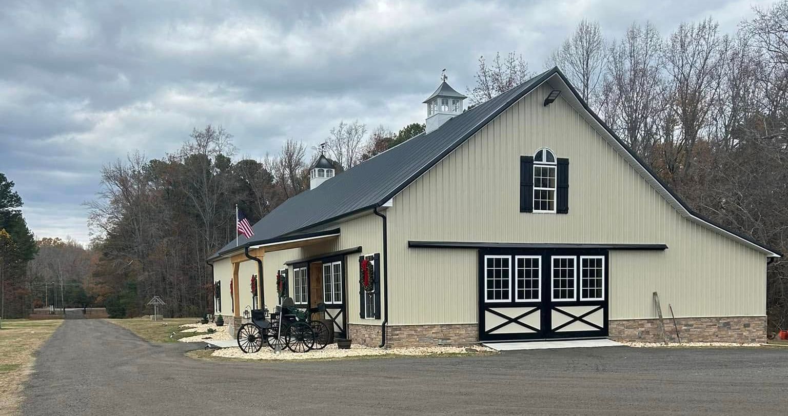 Cream-colored barn with black doors and roof, small cupola. Brick base. Overcast day, trees in background.