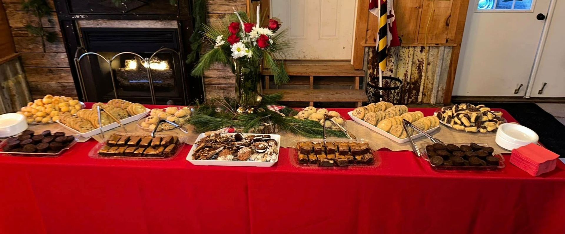 A table with a red tablecloth displays a variety of cookies and brownies, along with a floral arrangement.