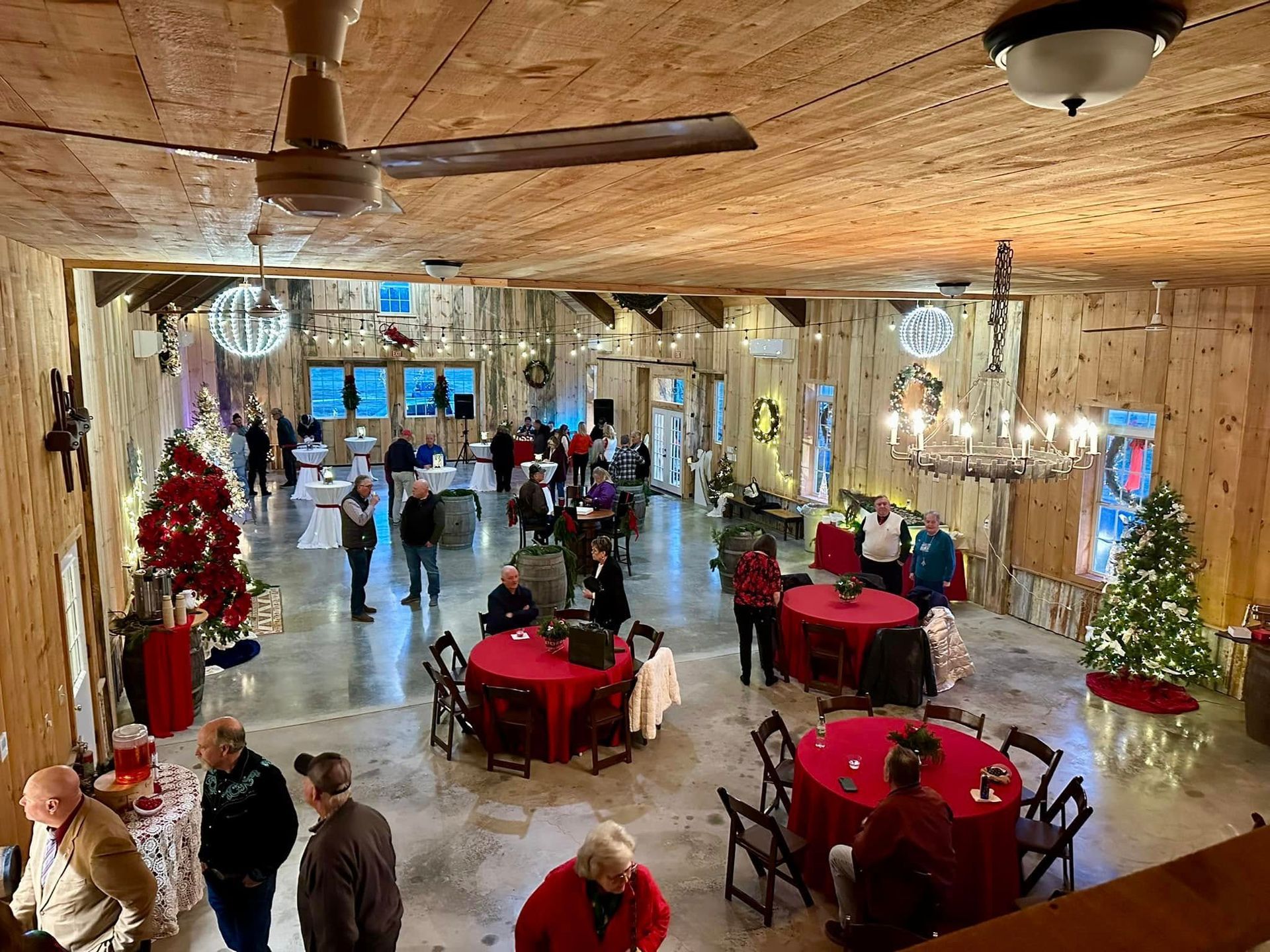 A large room with people at a holiday party; red tables, Christmas trees, wood paneling.