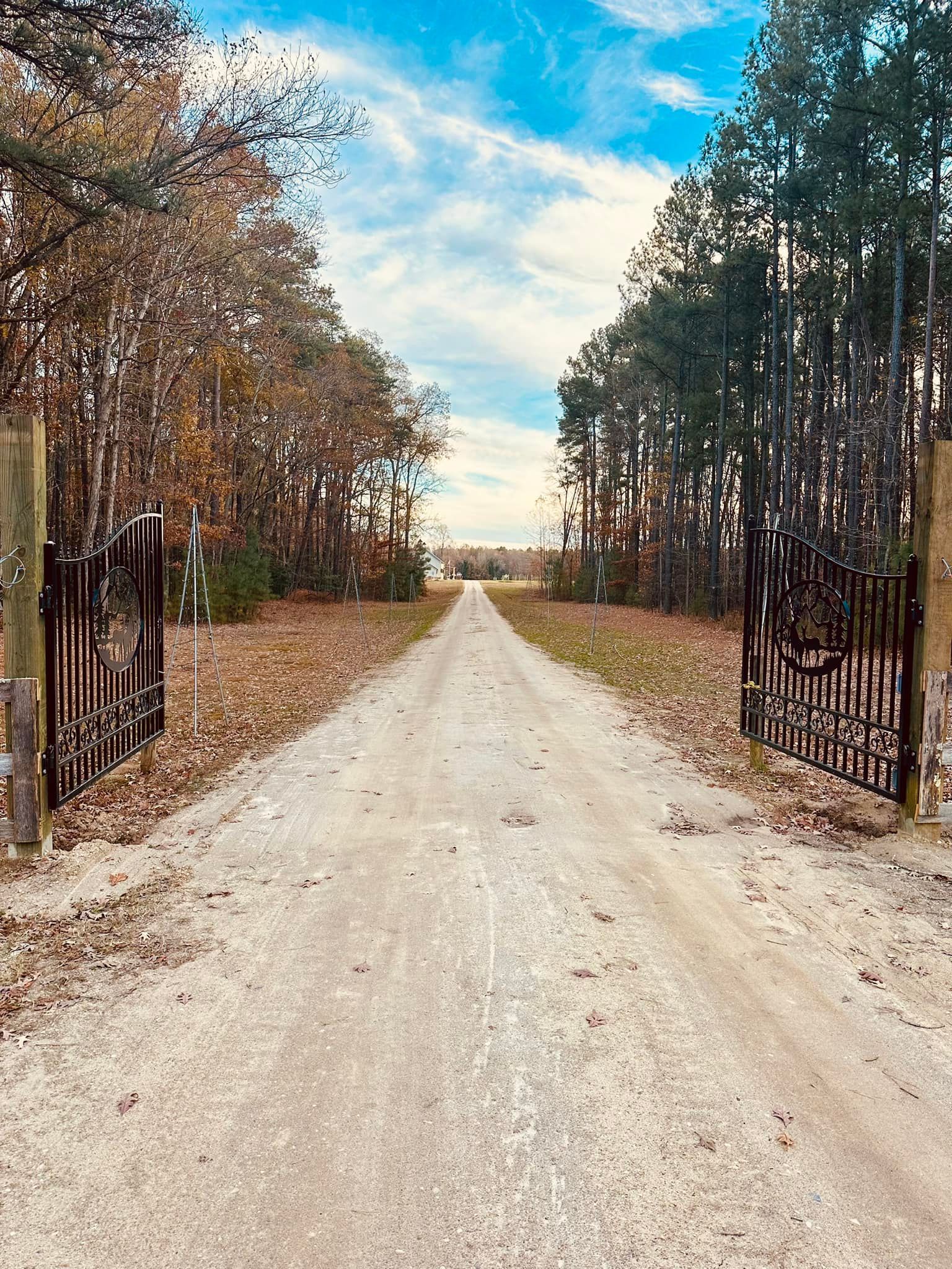 A long, gravel driveway leading to a house, framed by open black gates and fall trees under a blue sky.