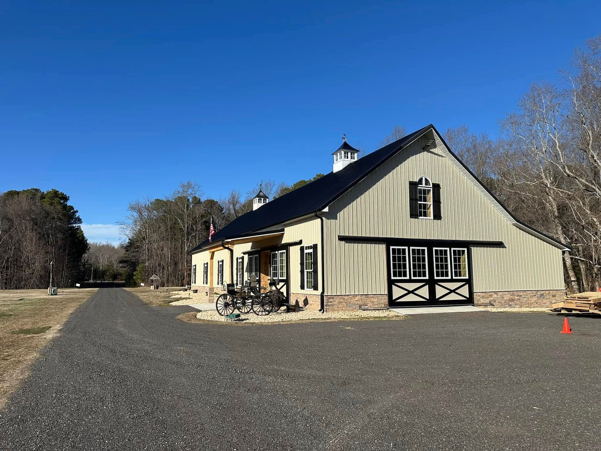 Tan barn with black doors, windows, and roof, on a gravel road under a blue sky.