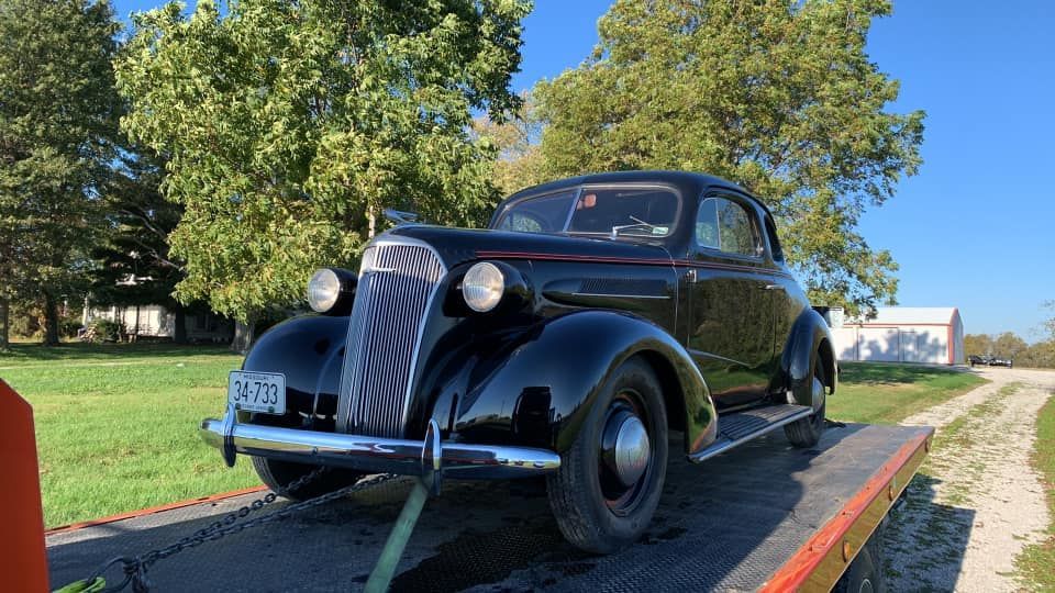 Black vintage coupe car on a tow truck, outdoors on a sunny day.