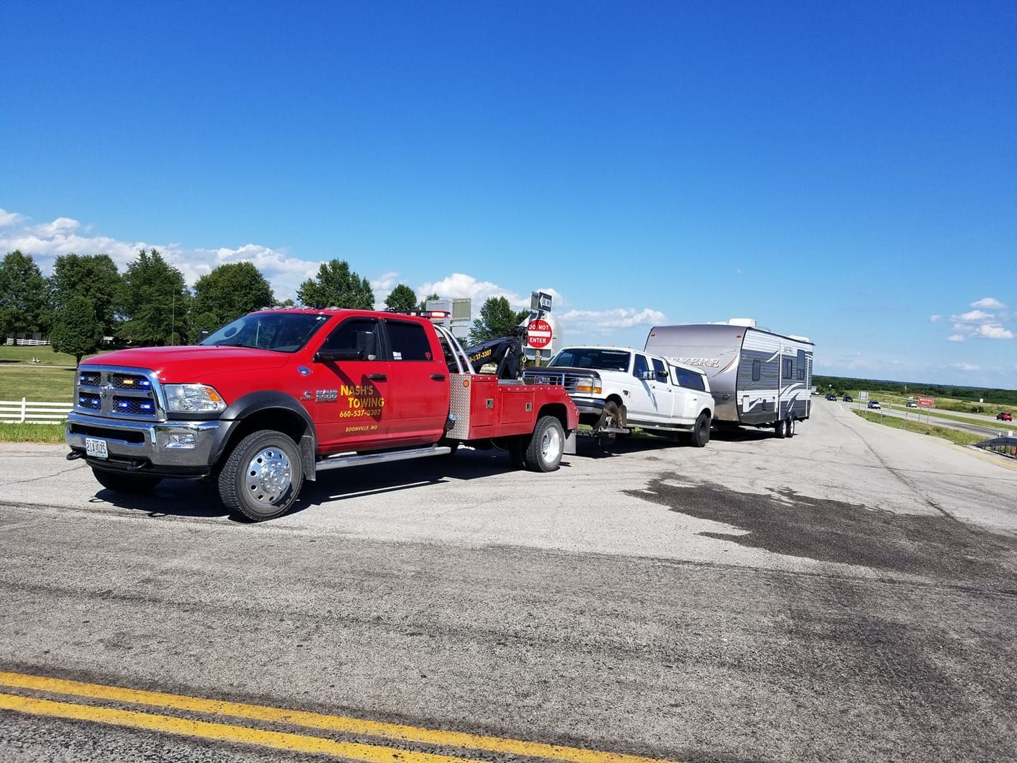 Red tow truck towing a white pickup truck and trailer on a road. Blue sky, sunny day.