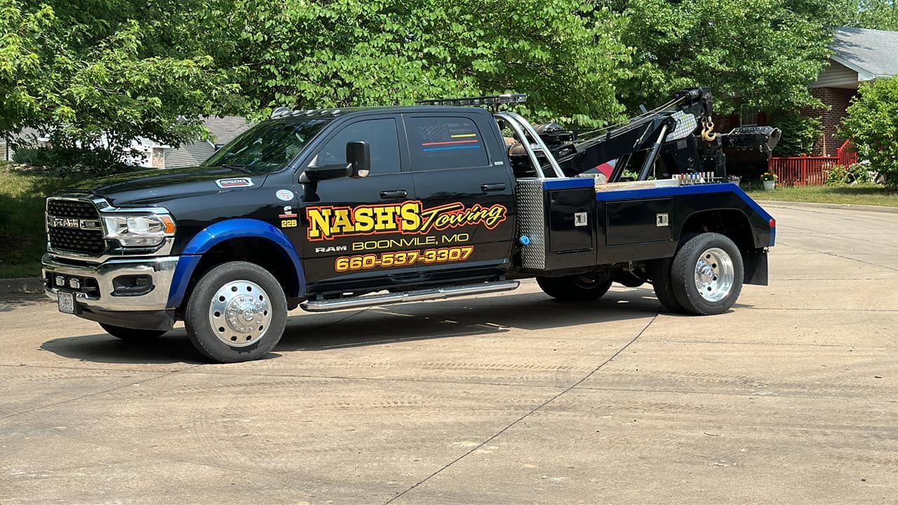 Black tow truck with Nash's Garage logo parked on pavement, blue and gold accents, trees in the background.