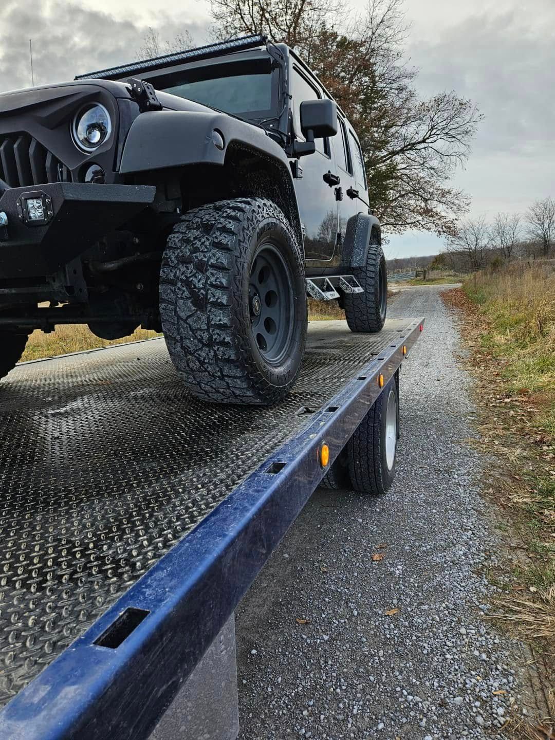 Black Jeep on a tow truck bed on a gravel road, overcast day.