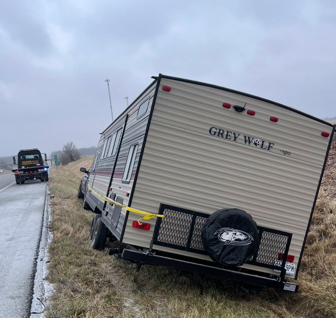 A Grey Wolf camper trailer partially off road, being towed away by a tow truck on a cloudy day.