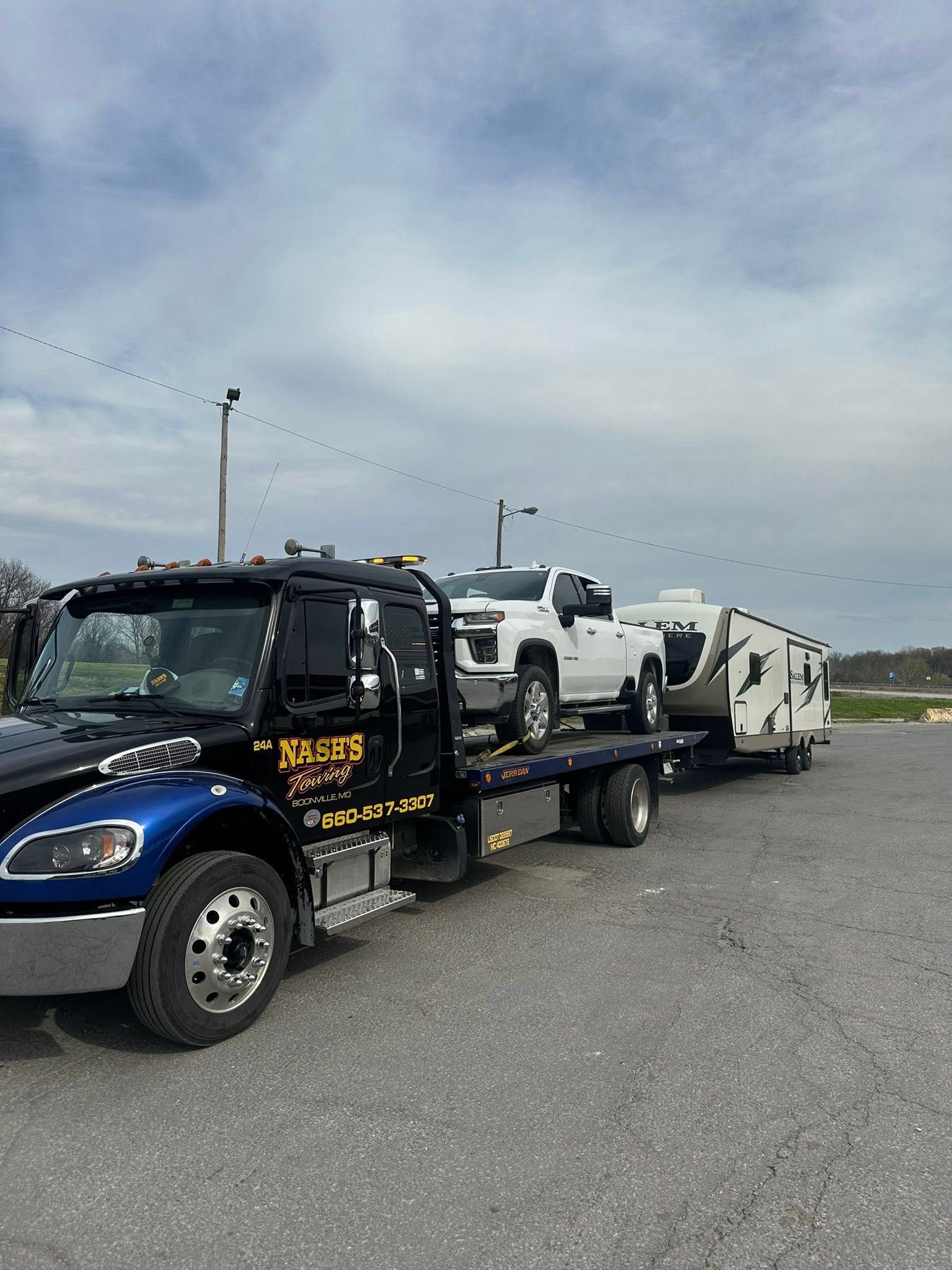 Tow truck hauling a white pickup truck with an attached camper trailer. Sky is cloudy.