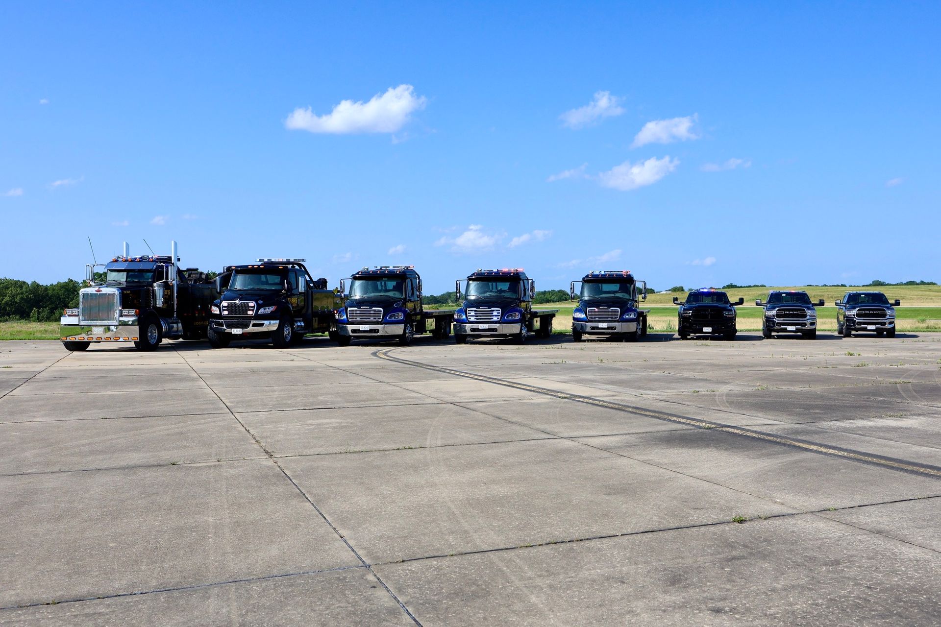 A line of tow trucks and pickups parked on a concrete surface under a blue sky.