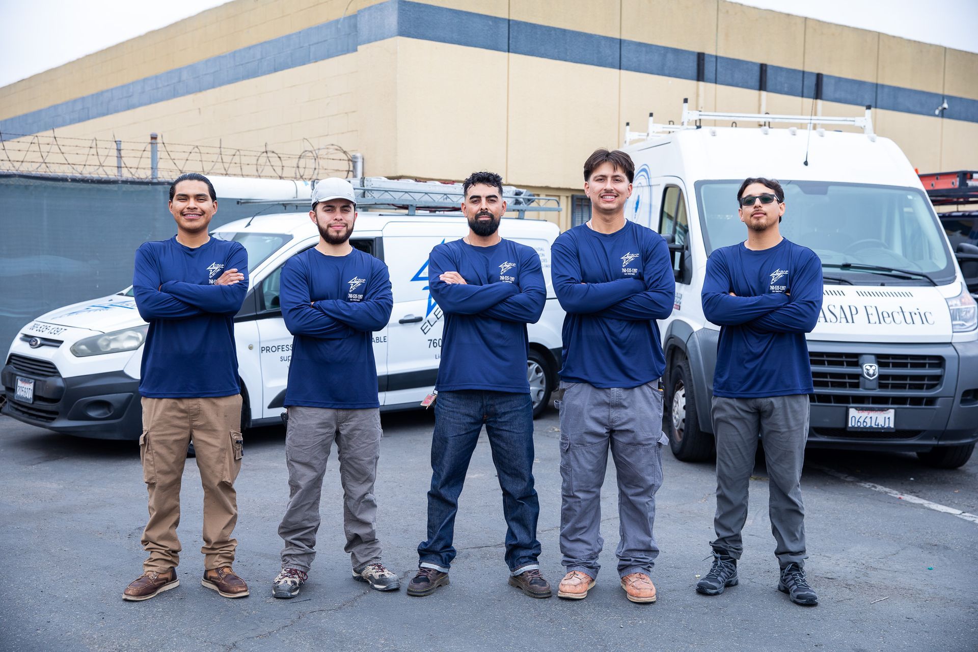 Five workers in blue shirts and work pants stand with arms crossed in front of white vans.