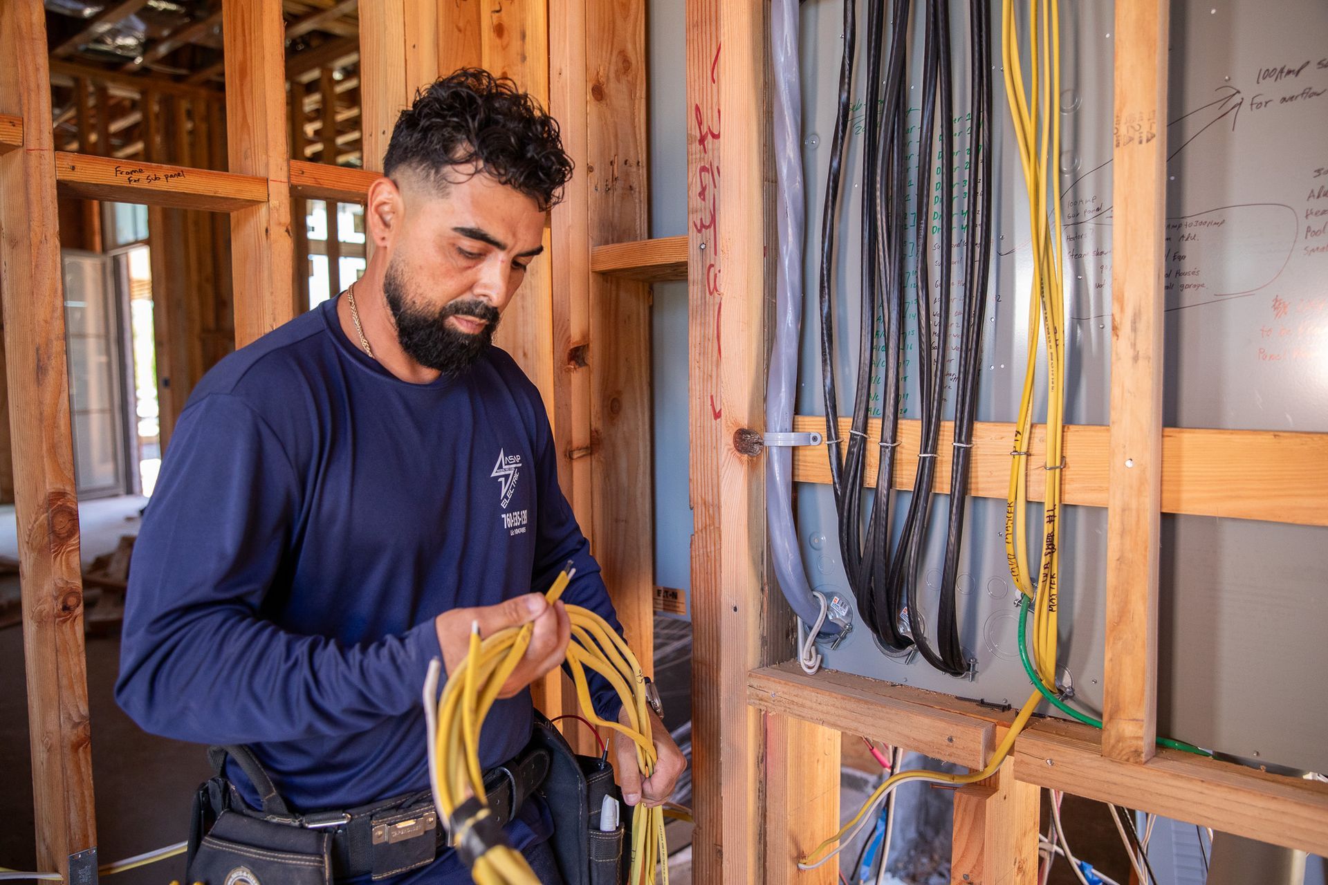 Electrician installing wiring in a wooden frame. Wears blue shirt, holds yellow wires. Interior construction site.
