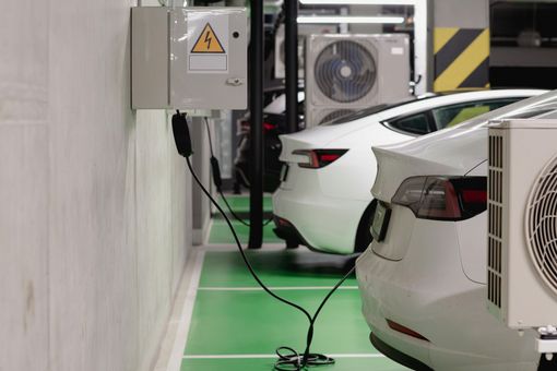 Two white electric cars charging in a parking garage, near electrical boxes and air conditioning units.