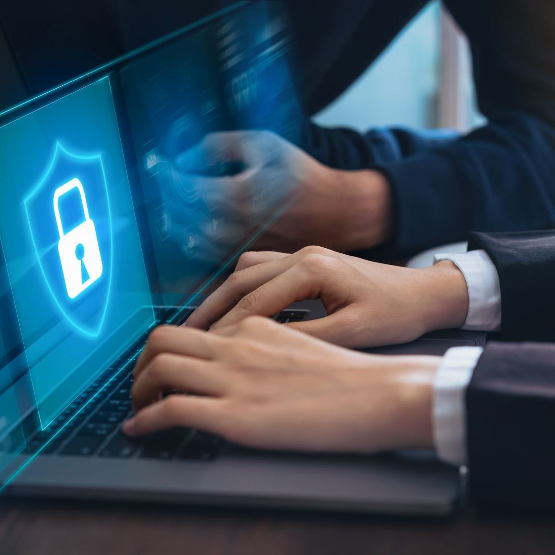 Hands typing on a laptop with a blue shield and padlock icon on the screen, representing cybersecurity.