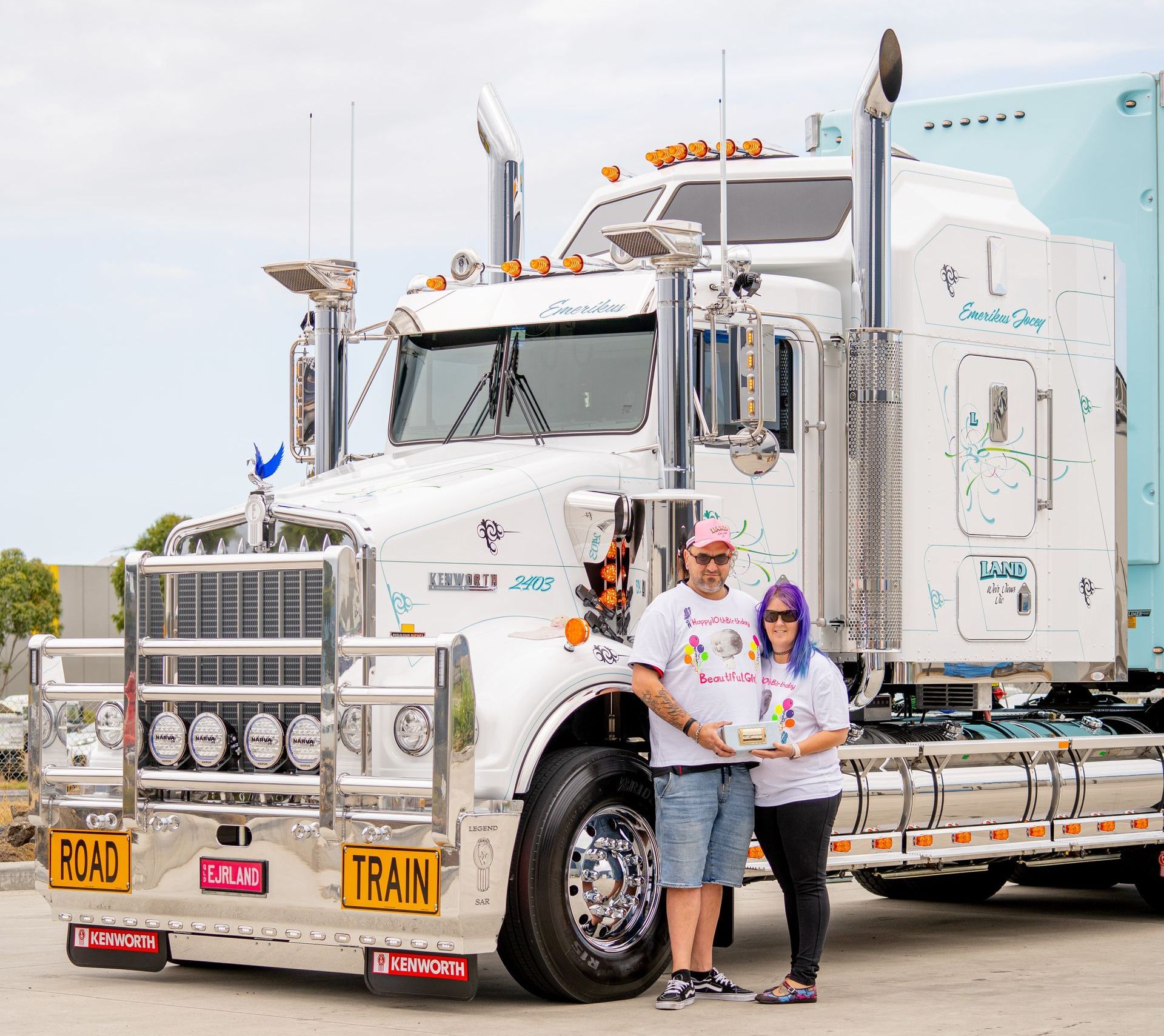 a man and a woman are standing in front of a semi truck that is decorated to support the Emerikus foundation. 