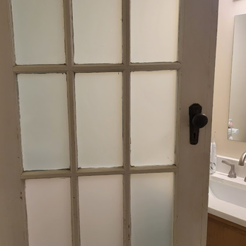 A light-colored door with nine frosted glass panes and a black door handle, partially obscuring a bathroom sink area.