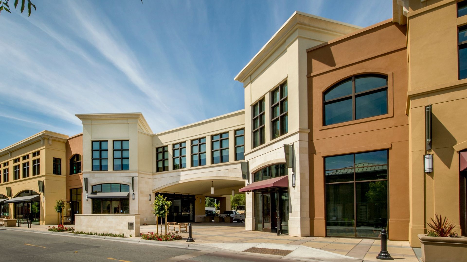 Modern multi-level commercial shopping plaza with beige and brown stucco facades, large windows, and a shaded walkway.