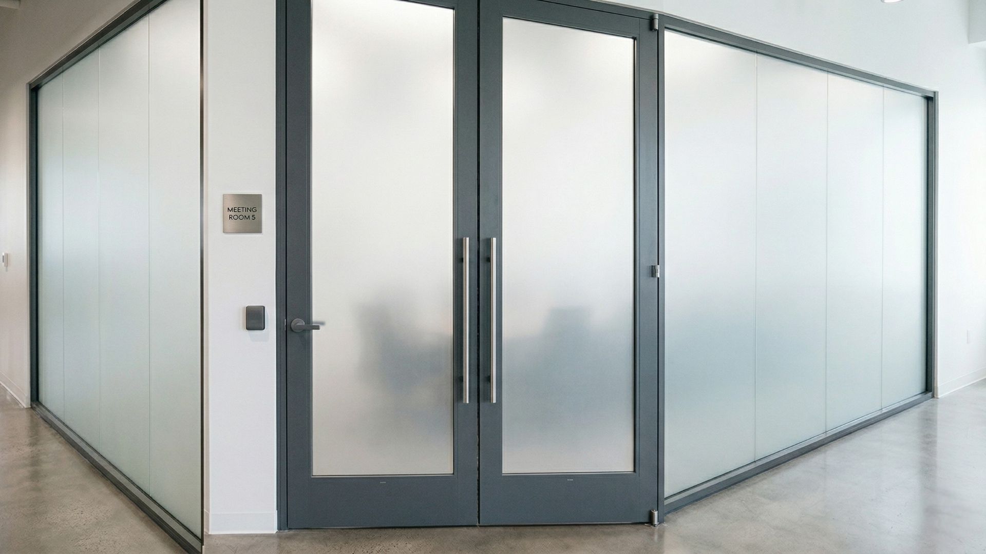 A modern office meeting room with dark gray frames, double glass doors, and frosted windows, set against white walls.