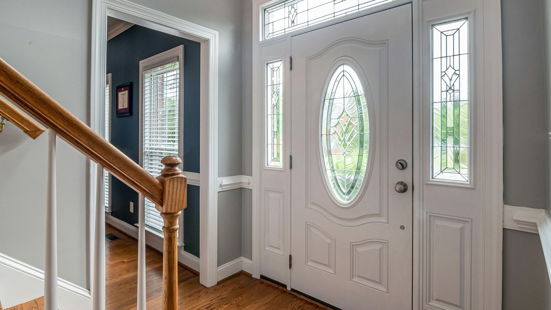 A bright entryway with a white front door featuring an oval glass insert and sidelights, next to a wooden staircase.