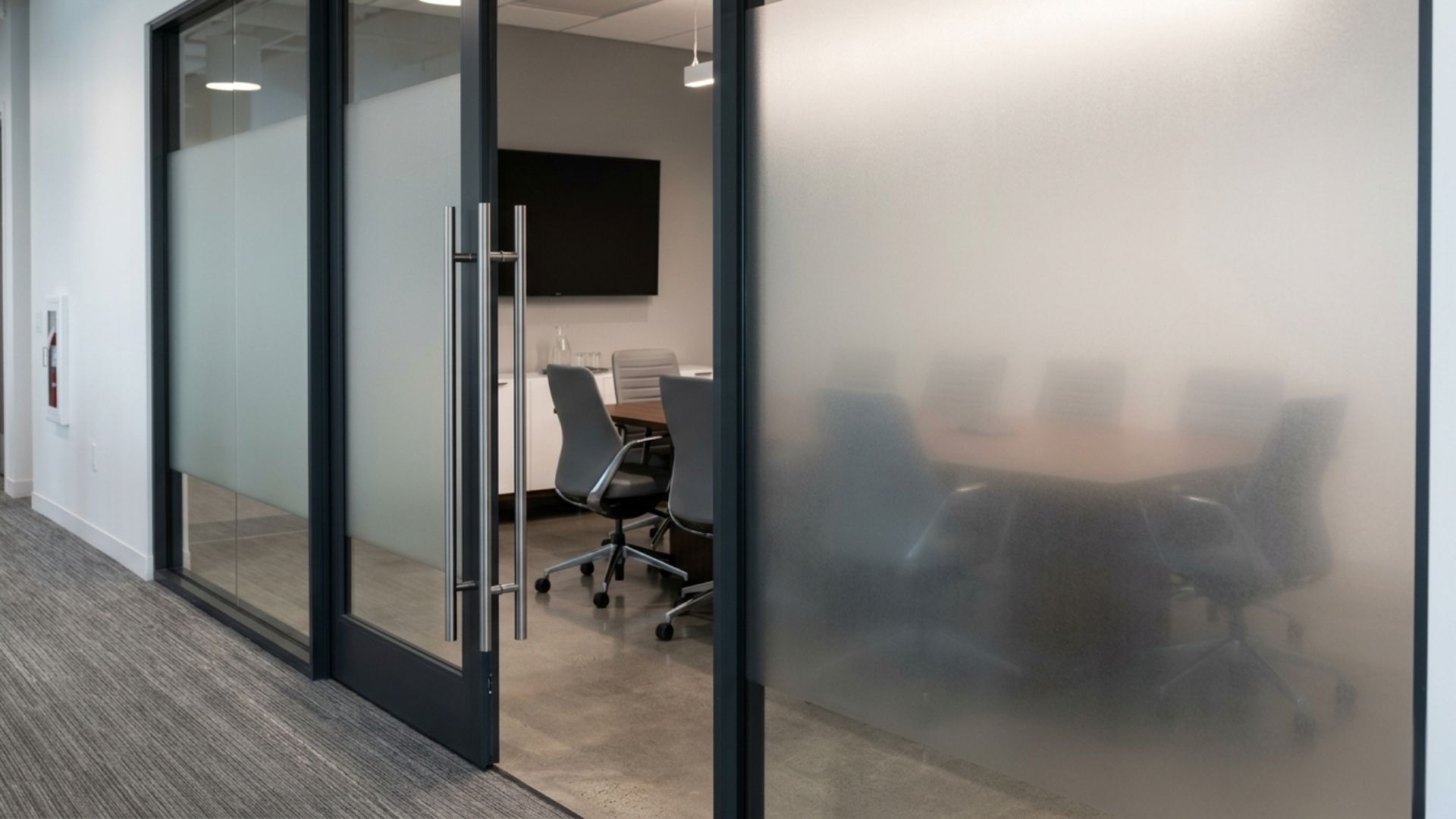 A modern glass-walled conference room with chairs, a table, and a screen inside, viewed from an office hallway.