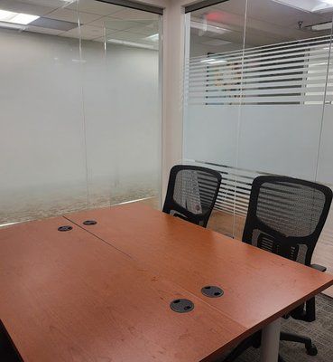 A wooden conference table with four cable ports and two black mesh office chairs in a modern, glass-walled office.