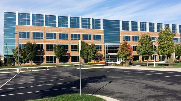 A modern four-story office building with brick walls and glass windows, seen from a parking lot on a sunny day.