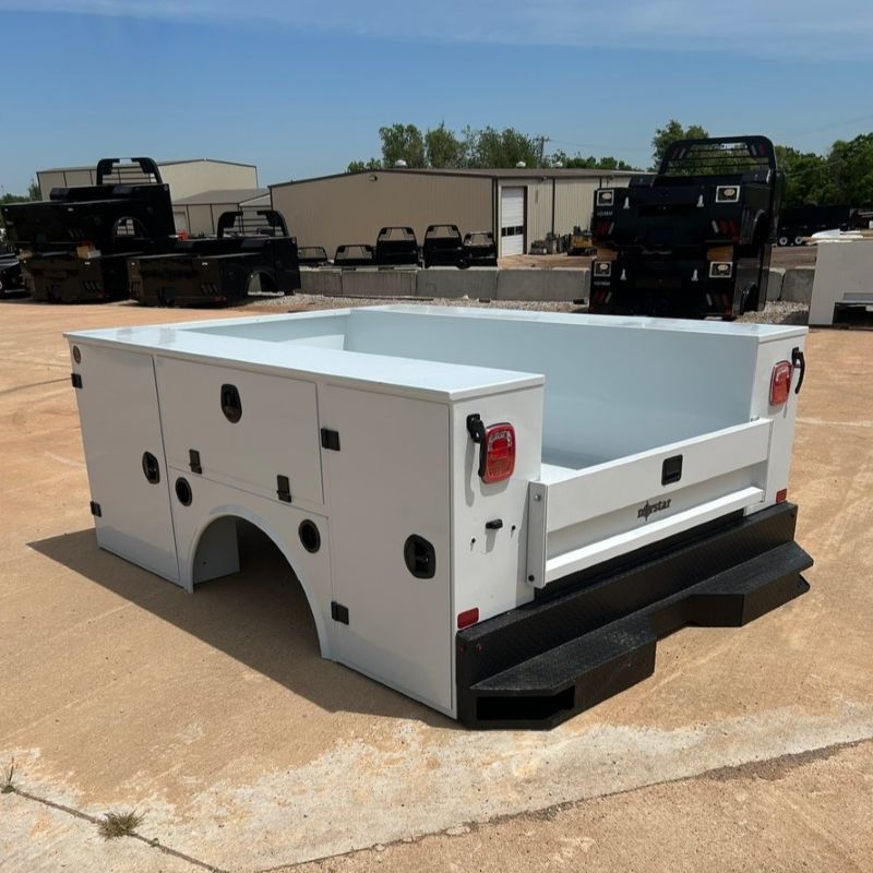 White utility truck bed body with black step bumper, shown outdoors on a lot with trucks in the background
