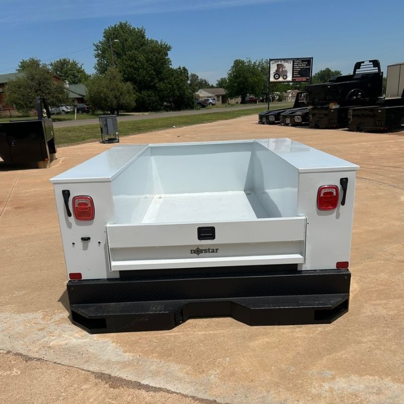 White utility trailer with open bed and black bumper, parked outdoors on a concrete lot.