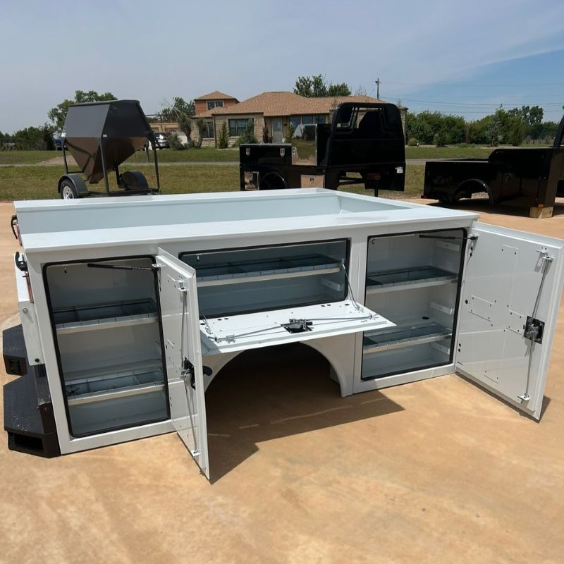 White utility bed with open doors and drawers on a driveway, showing a built-in storage.