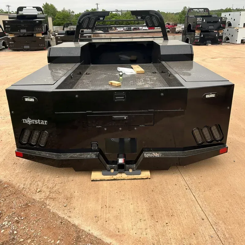 Black flatbed truck bed with tool compartments parked on a dirt lot, other trucks in the background