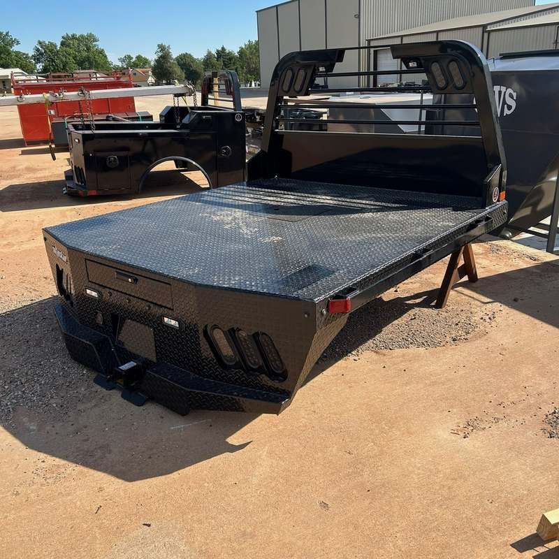 Black flatbed truck with diamond-plate steel deck parked on a dirt lot, with another truck behind it.