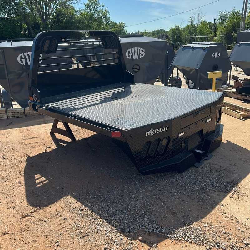 Black flatbed utility trailer with head rack and diamond plate deck in a dirt lot outside a warehouse.
