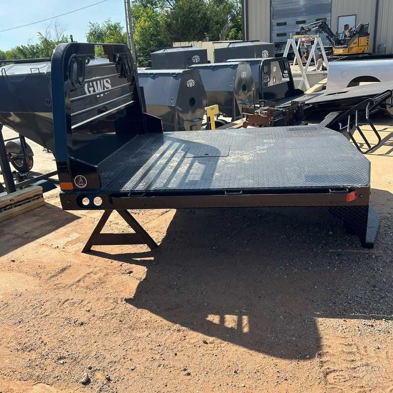 Black flatbed truck bed with ramps in a dusty outdoor lot, with equipment and trailers in the background