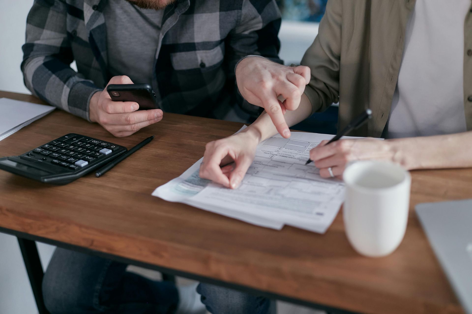 Couple reviewing documents together at a wooden table. One points to a paper while the other holds a phone.
