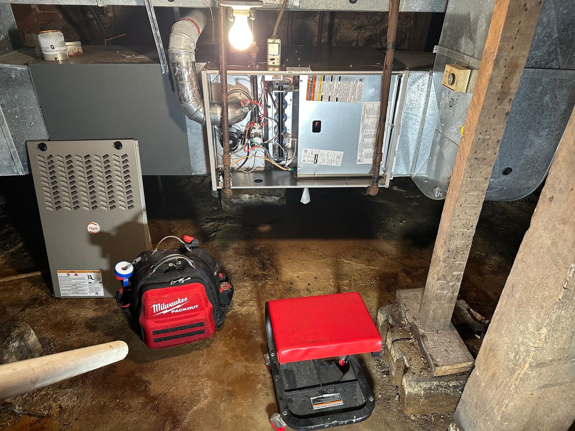 An open HVAC furnace unit in a dimly lit basement with a red tool bag and a red rolling stool on the floor.