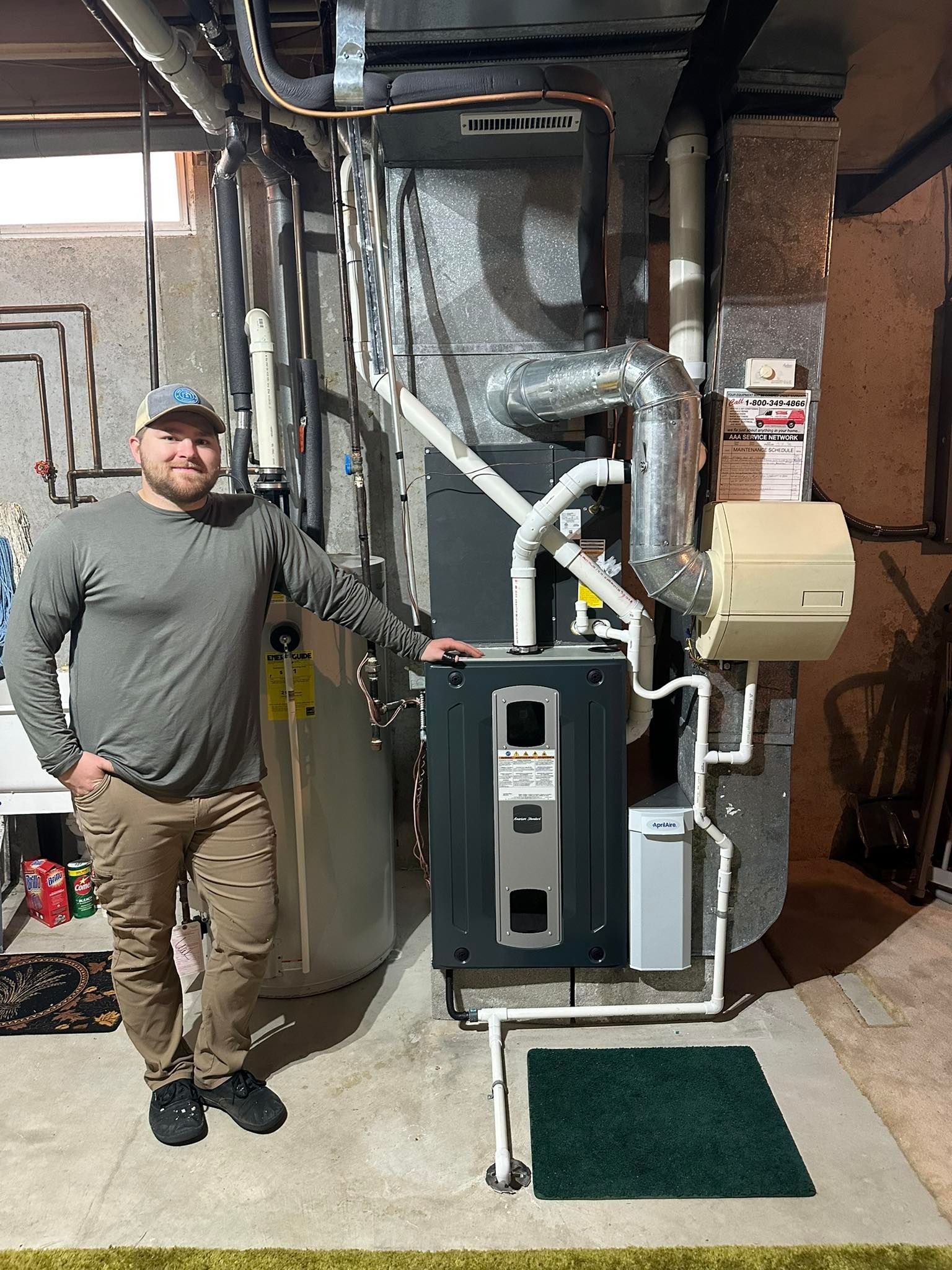 A person stands next to a newly installed residential HVAC furnace and water heater system in a basement setting.