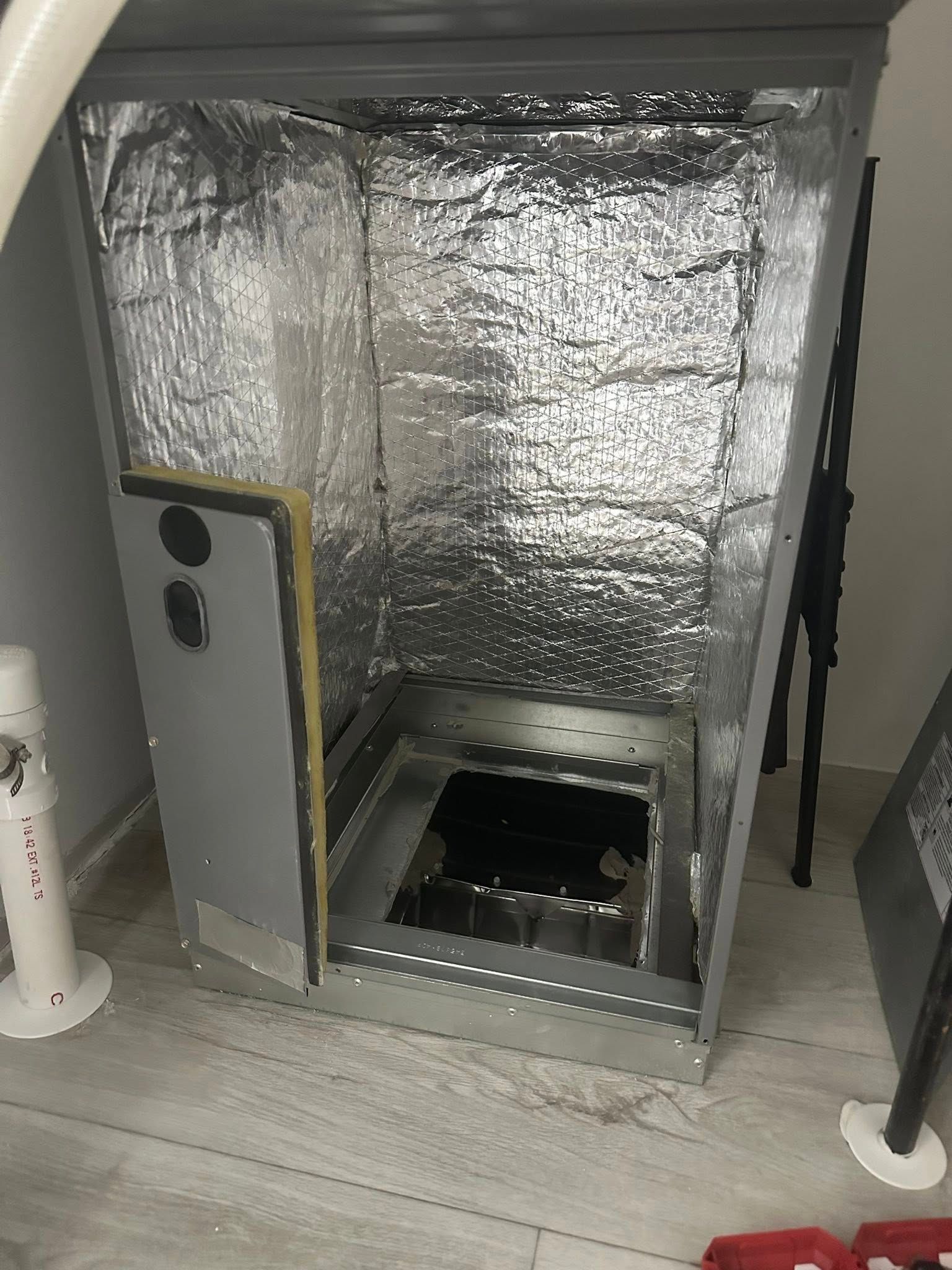 An open, silver-lined HVAC furnace cabinet sitting on a wooden floor in a room.