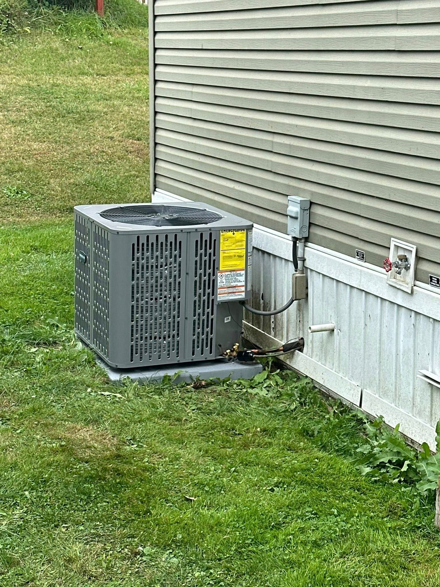 A grey air conditioning unit sits on a concrete pad next to the light green siding and white skirting of a house.