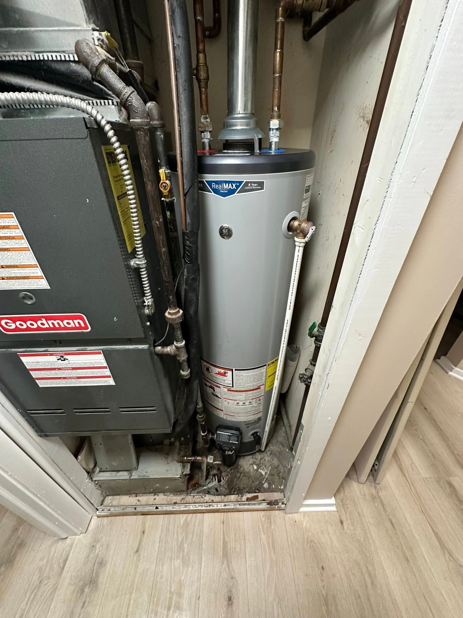 A tall, grey gas water heater stands beside a Goodman furnace in a narrow utility closet.