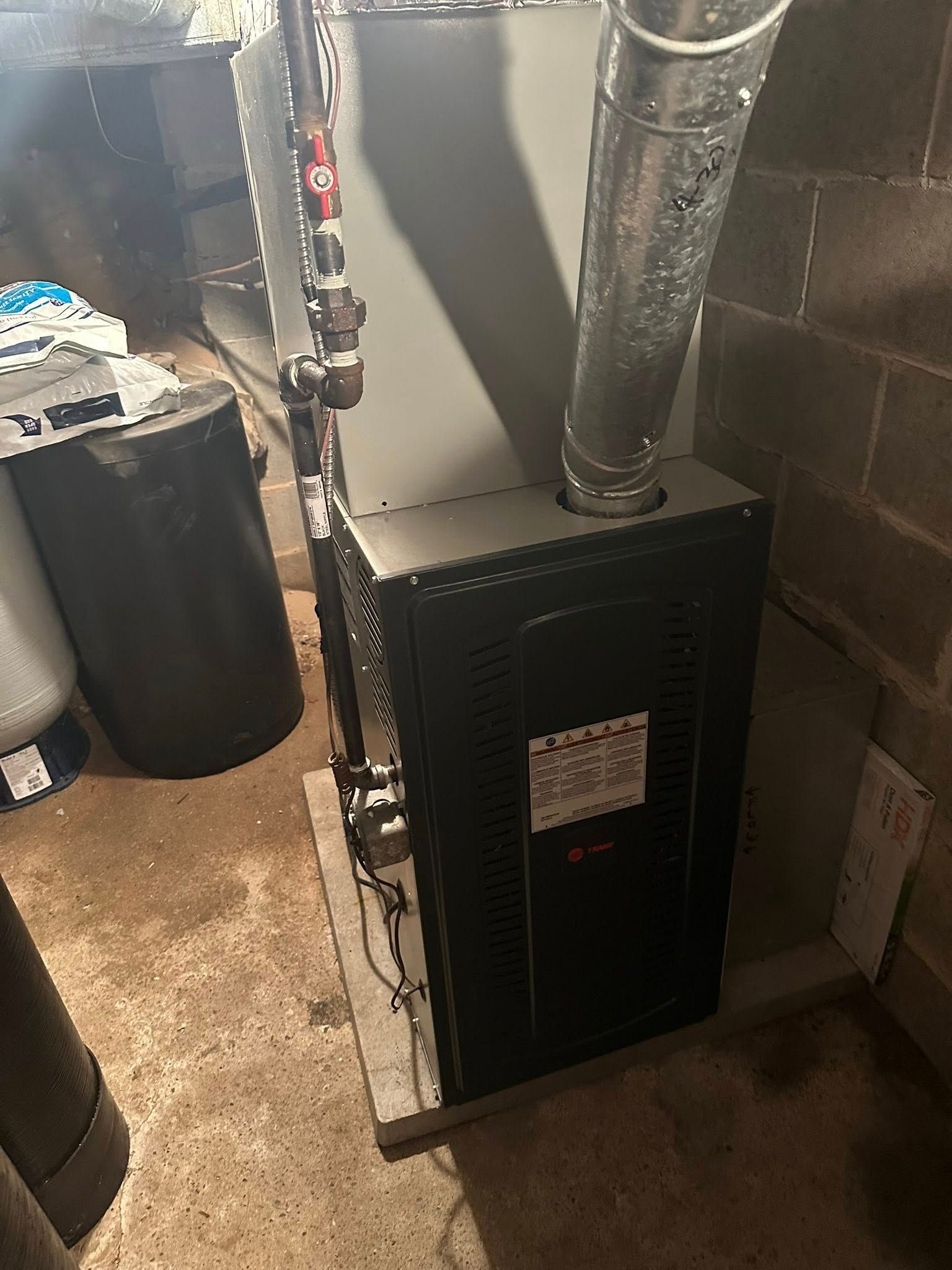 A black and gray natural gas furnace stands in a basement with a silver metal flue pipe attached to the top.