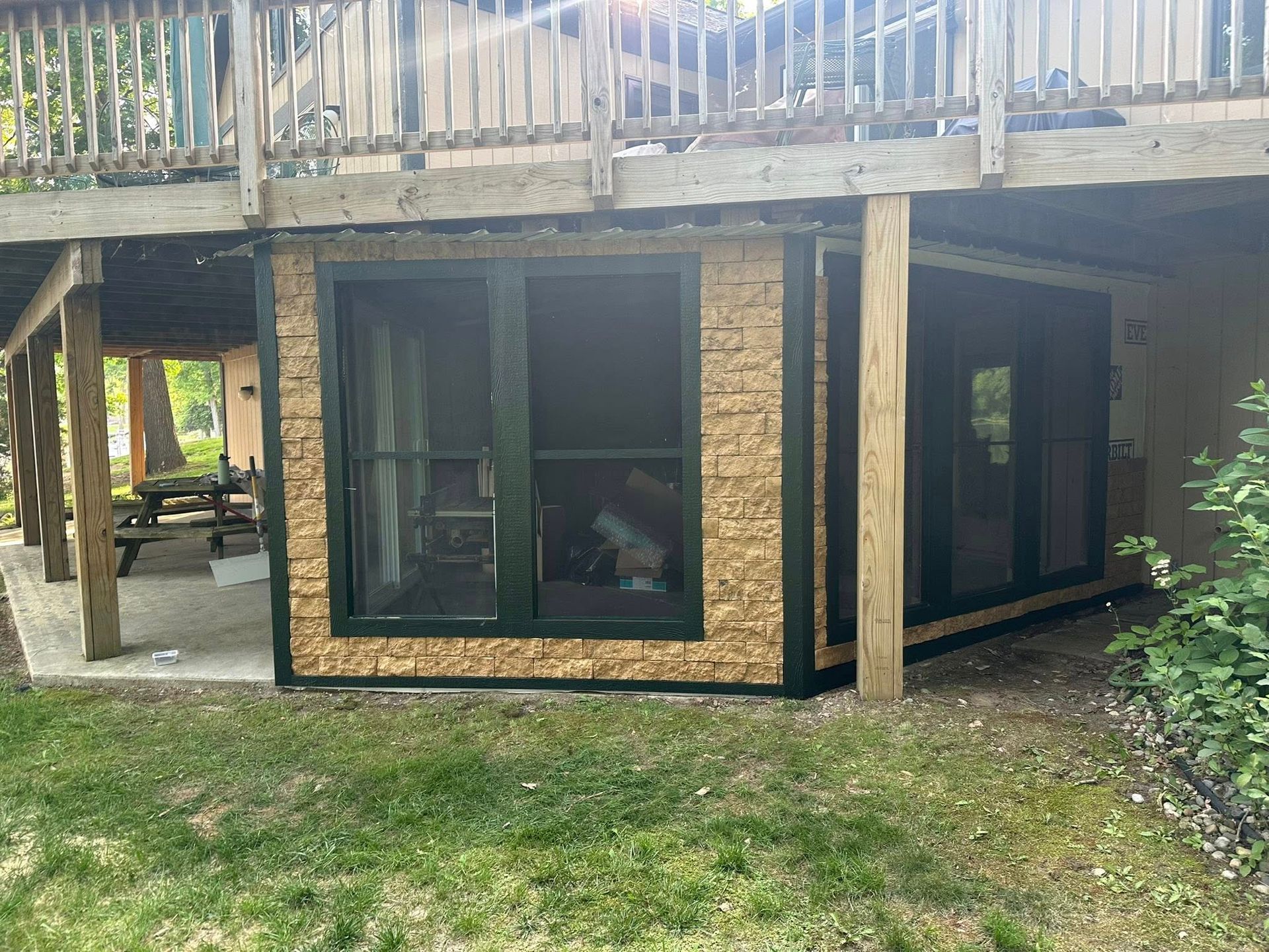 A house deck with a partially finished enclosed room beneath it featuring dark-framed windows and OSB exterior walls.