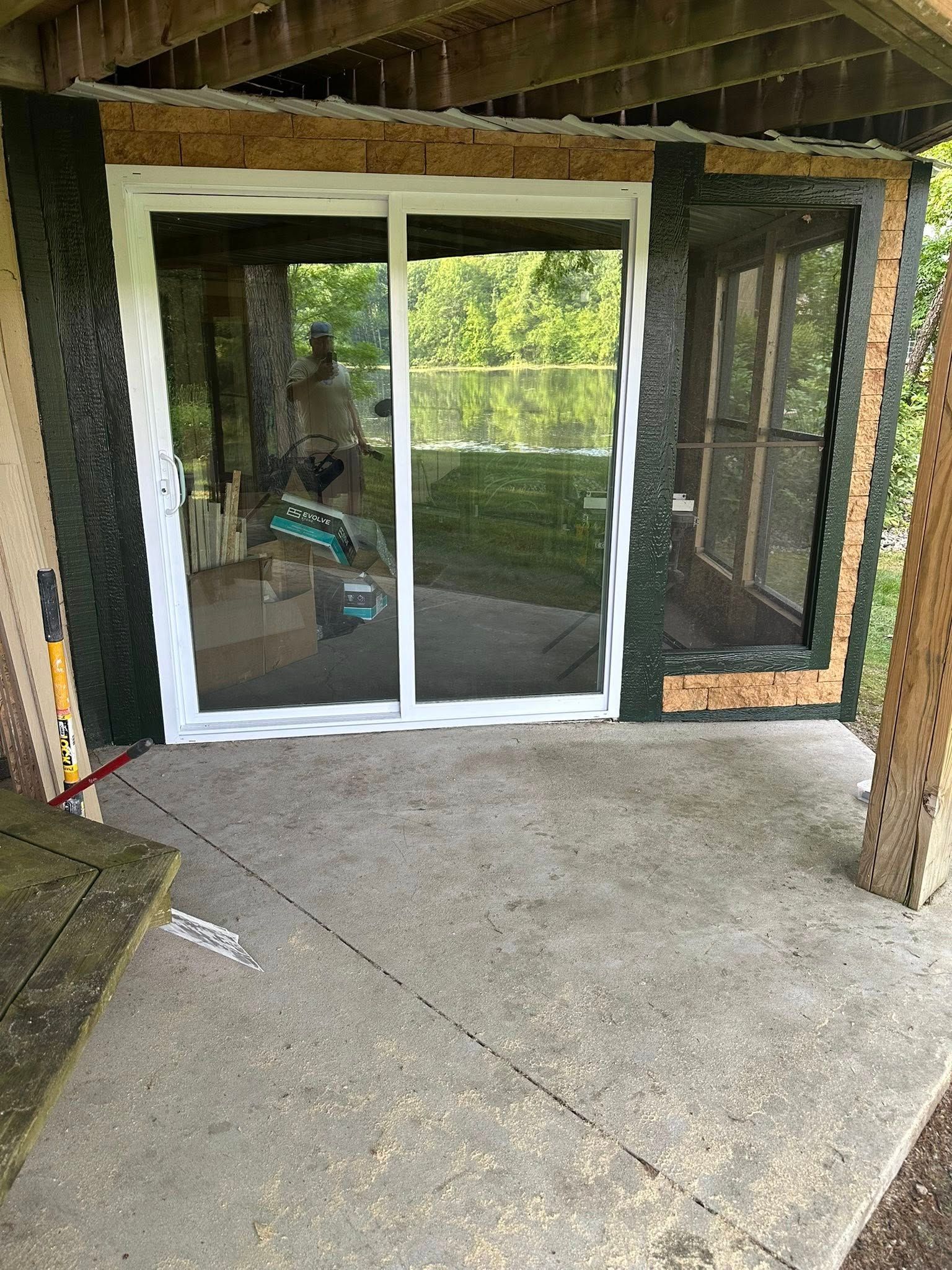 A white sliding glass door installed in an unfinished wooden frame on a concrete patio overlooking a pond.