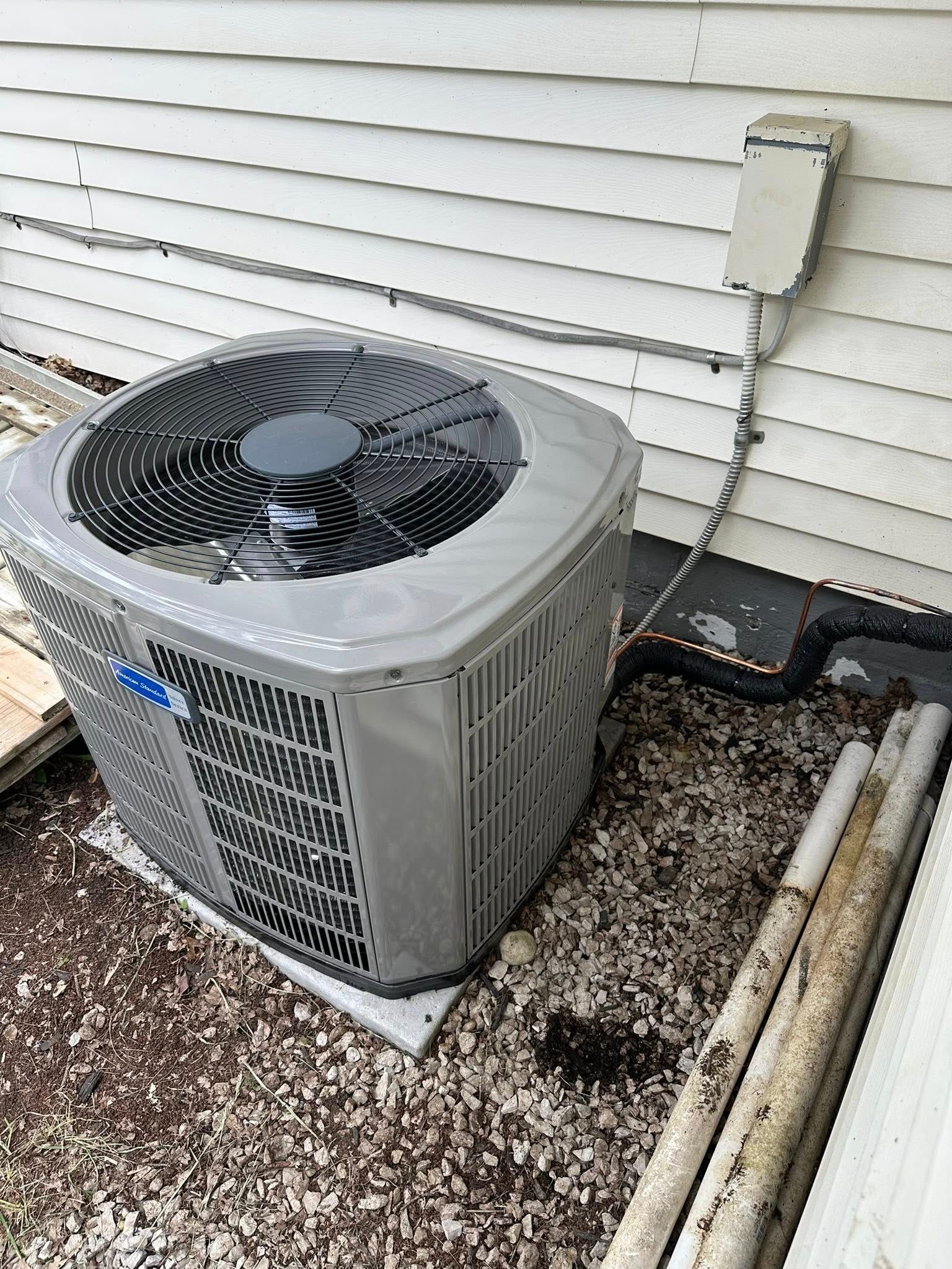 A gray residential outdoor air conditioning unit sits on a gravel patch against the white siding of a house.