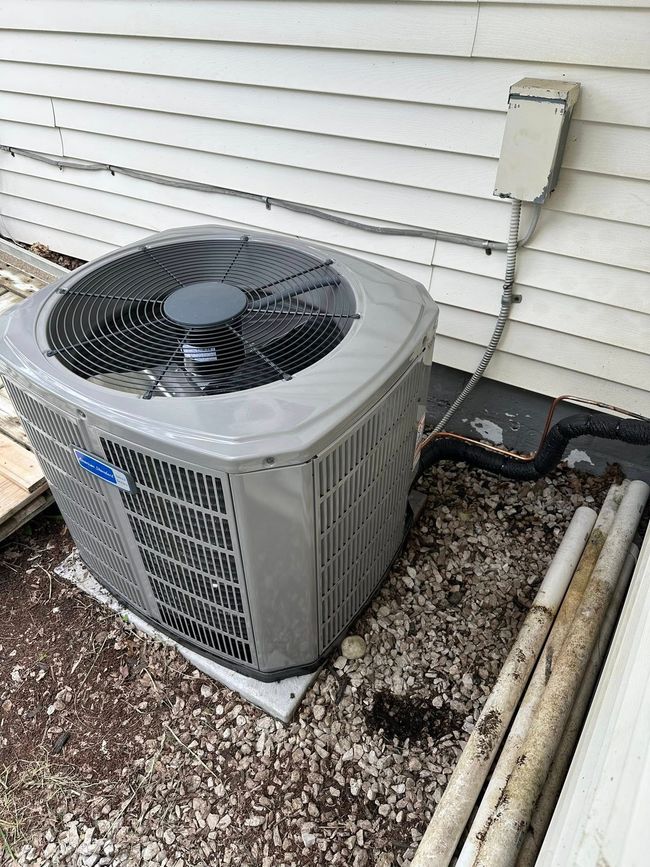 A gray HVAC unit sits outside a house with brown horizontal siding, near a brick foundation and some garden greenery.