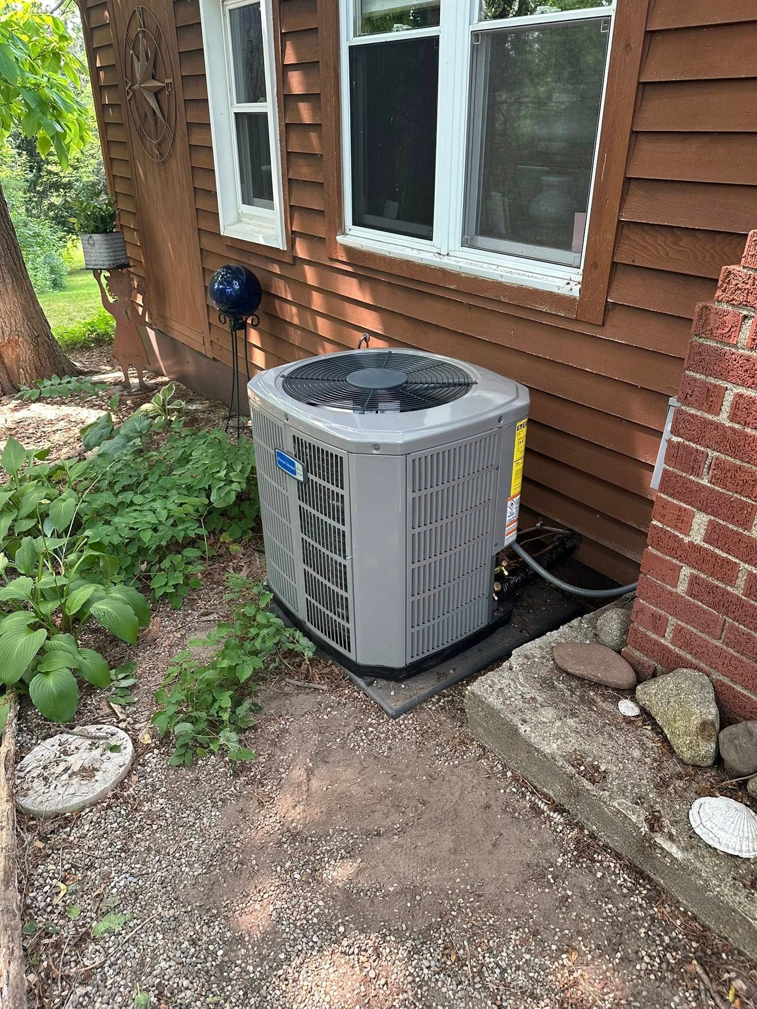 A gray air conditioning unit sits on a concrete pad outside a brown house next to a brick wall and green garden foliage.
