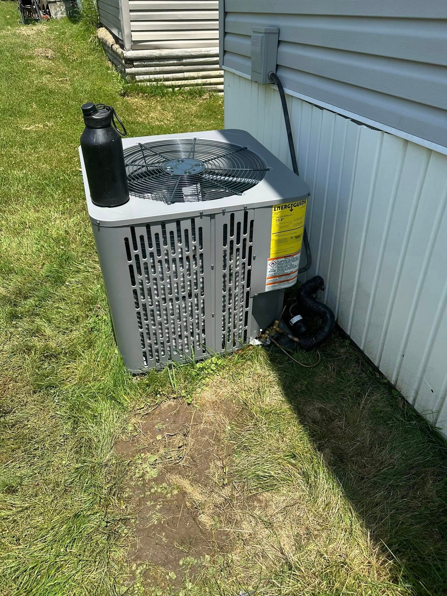 A gray air conditioning unit sits on grass next to the white siding of a house, with a black water bottle on top.