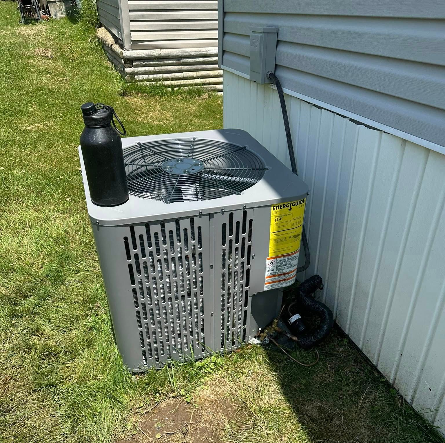 A gray central air conditioning unit sits on grass next to the side of a light-colored house with a black bottle on top.