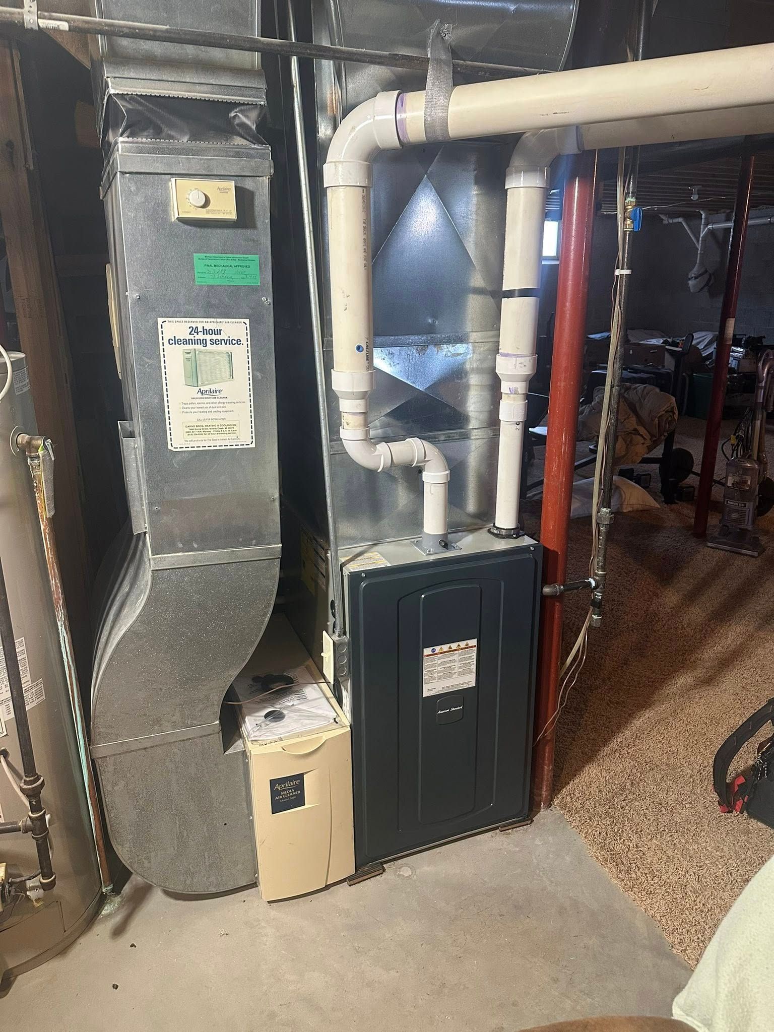 A basement furnace system with metal ductwork, white PVC venting pipes, and a grey service panel on a concrete floor.