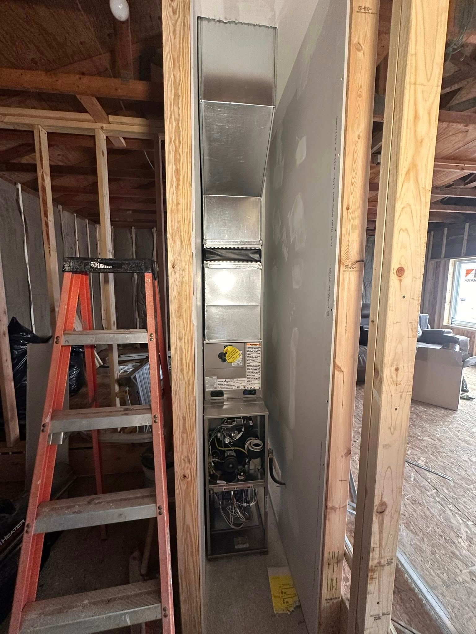 An HVAC unit installed inside a narrow wall framing cavity next to an orange stepladder in an unfinished room.