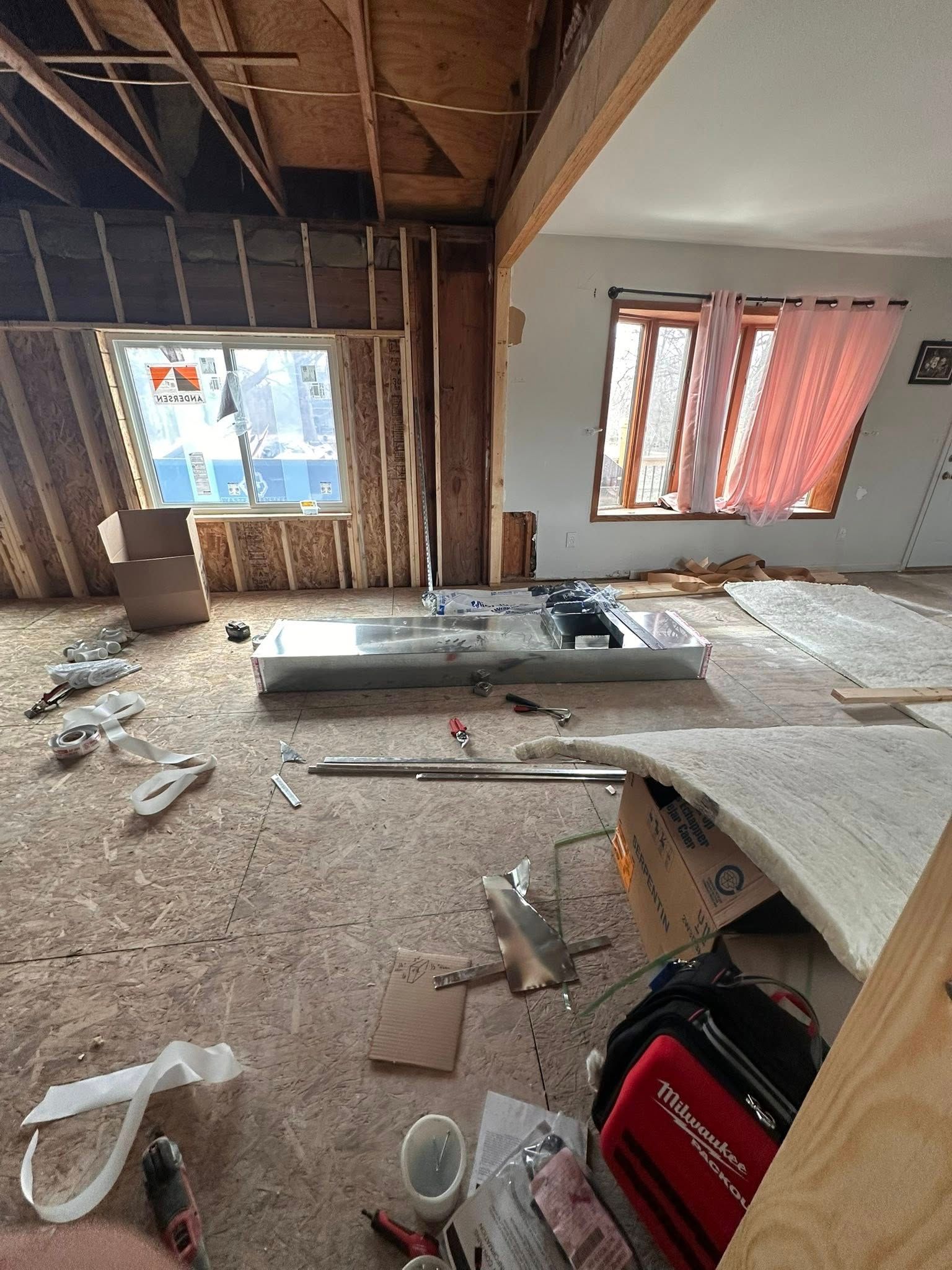 An interior view of a room under construction, showing exposed framing, a floor-level air duct, and building supplies.