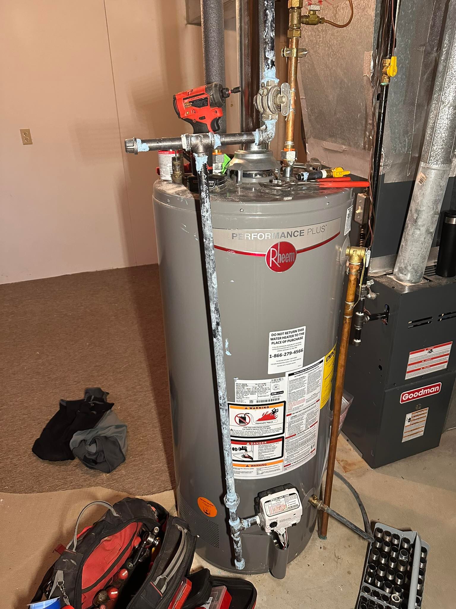 A grey Rheem water heater in a basement with plumbing pipes connected to the top and a tool bag on the floor nearby.