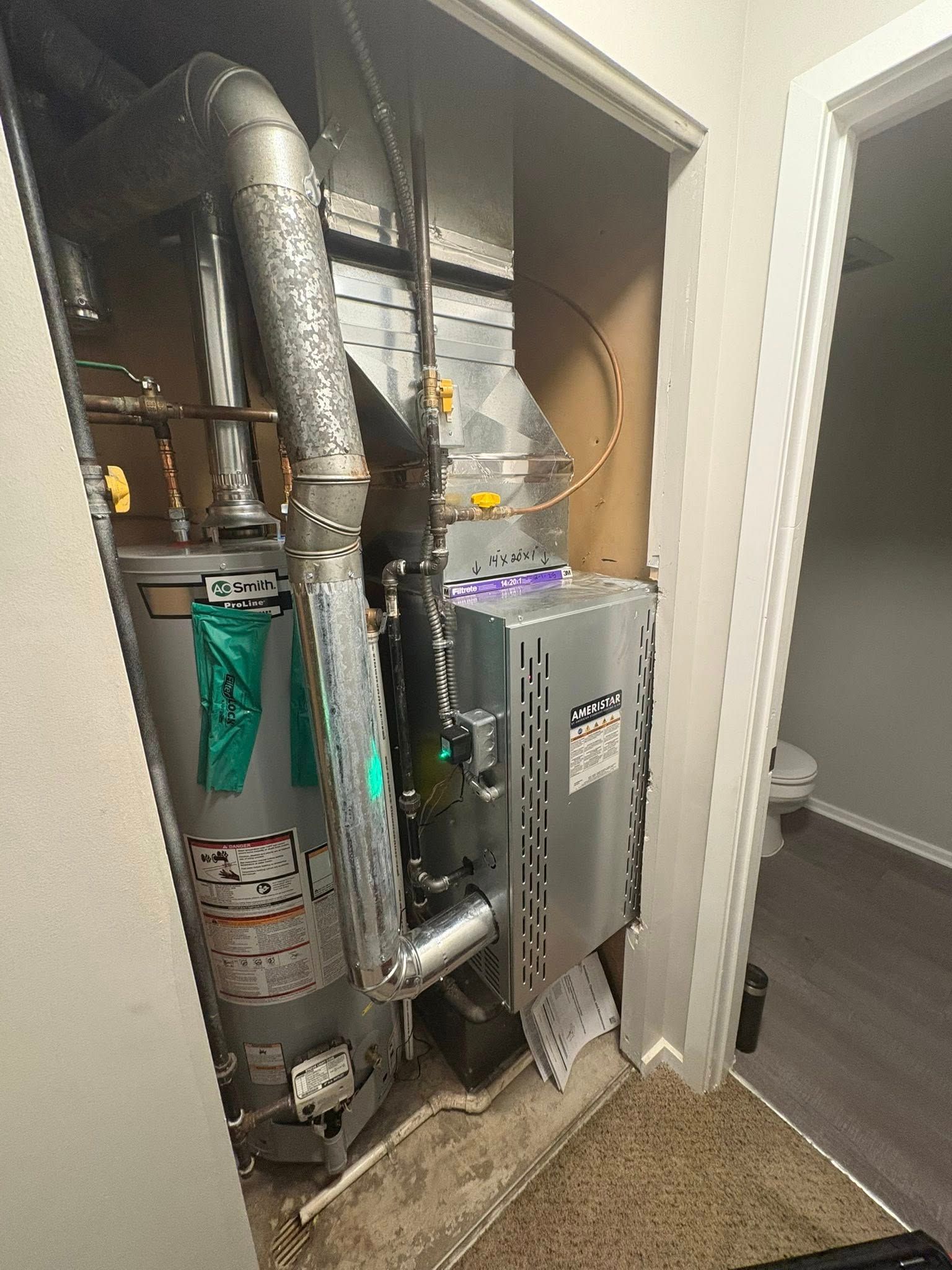 A water heater sits next to a metal HVAC unit inside a utility closet, with ductwork running above and behind them.